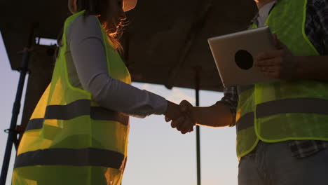Builder-man-with-a-tablet-and-a-woman-inspector-in-white-helmets-shake-hands-at-sunset-standing-on-the-roof-of-the-building.-Symbol-of-agreement-of-successful-work